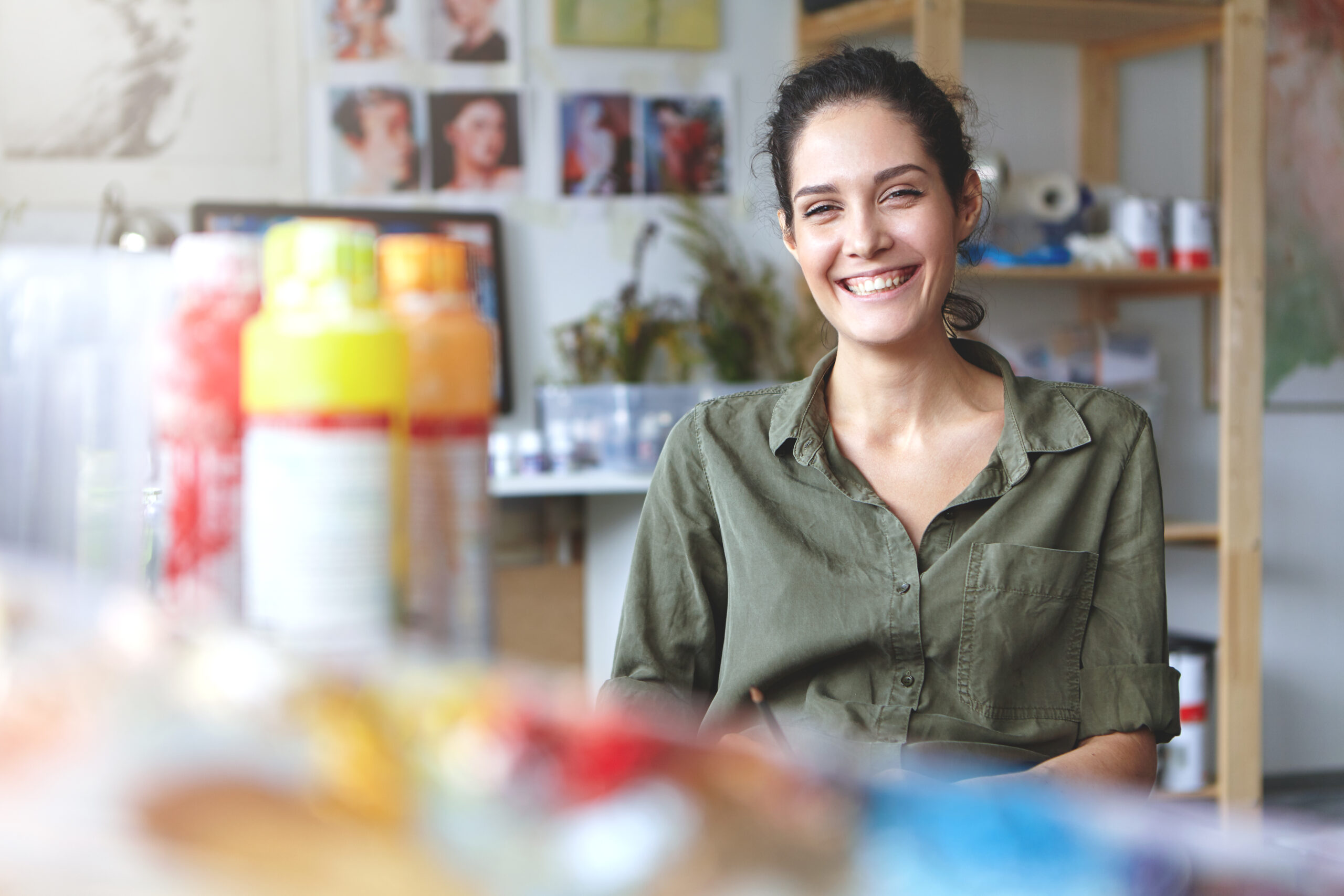 Picture of charming charismatic young female artist wearing khaki shirt grinning broadly feeling happy about her job and creating process, sitting at workshop, surrounded with painting accessories